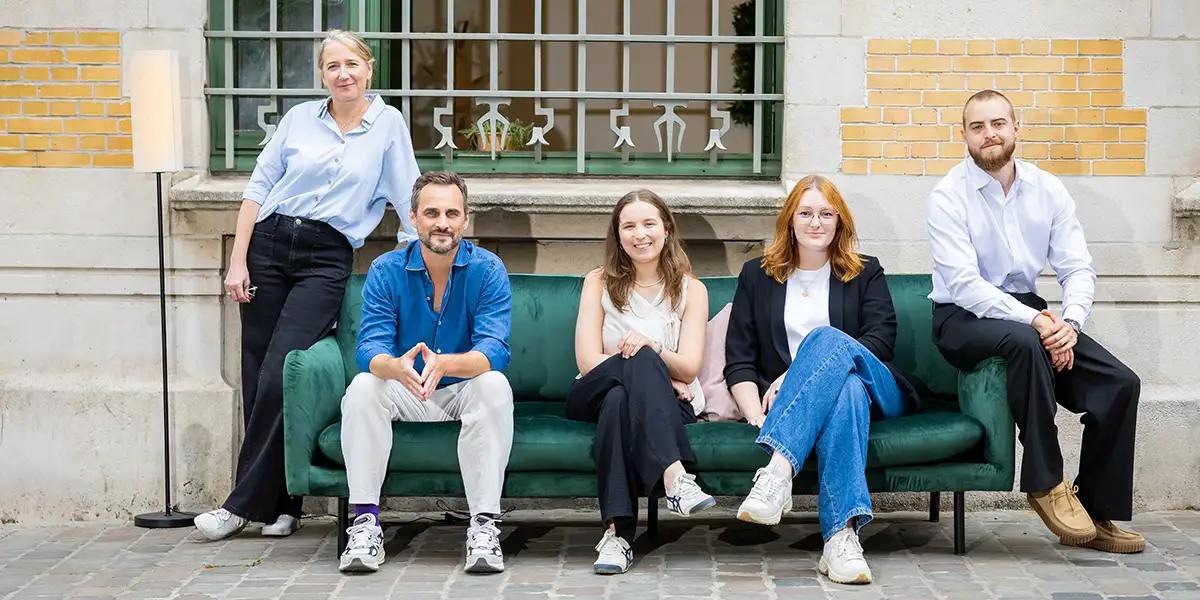 Cinq personnes posent sur un canapé vert devant un mur en briques jaunes et blanches, avec une fenêtre à barreaux.