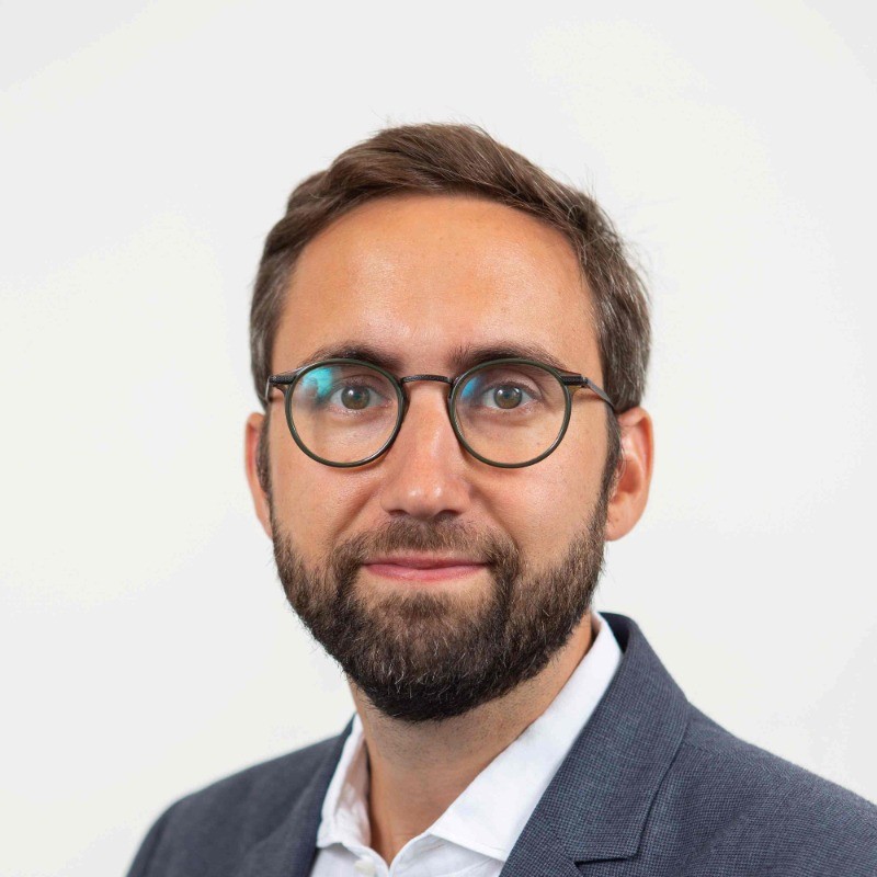 Un homme portant des lunettes et une barbe, vêtu d'une veste grise et d'une chemise blanche, sur fond clair.