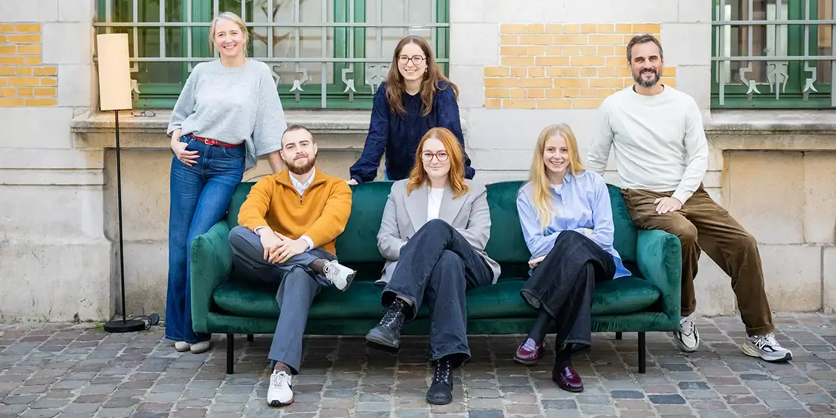 Un groupe de six personnes posent devant un bâtiment en briques, deux assises sur un canapé vert et quatre debout.