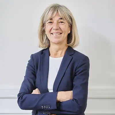 Une personne souriante aux cheveux gris porte un costume bleu et une chemise blanche, posant devant un mur blanc.