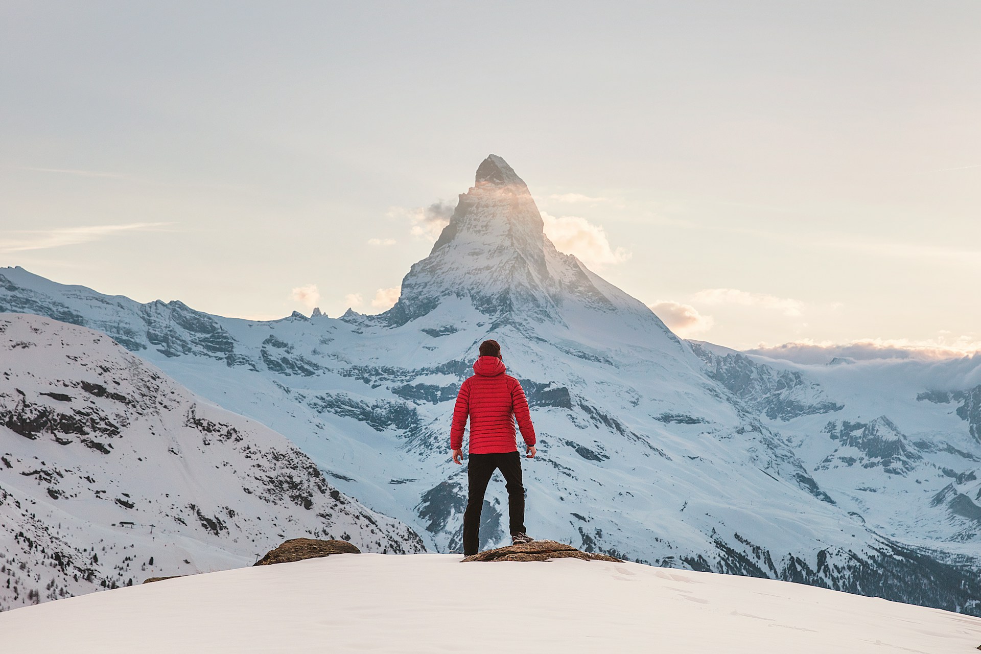 Une personne en veste rouge se tient sur une colline enneigée, face à une montagne majestueuse sous un ciel clair.