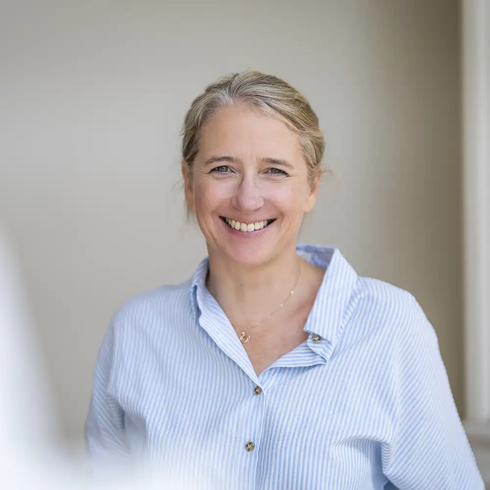Une personne souriante porte une chemise à rayures bleues et blanches, posant devant un mur beige.
