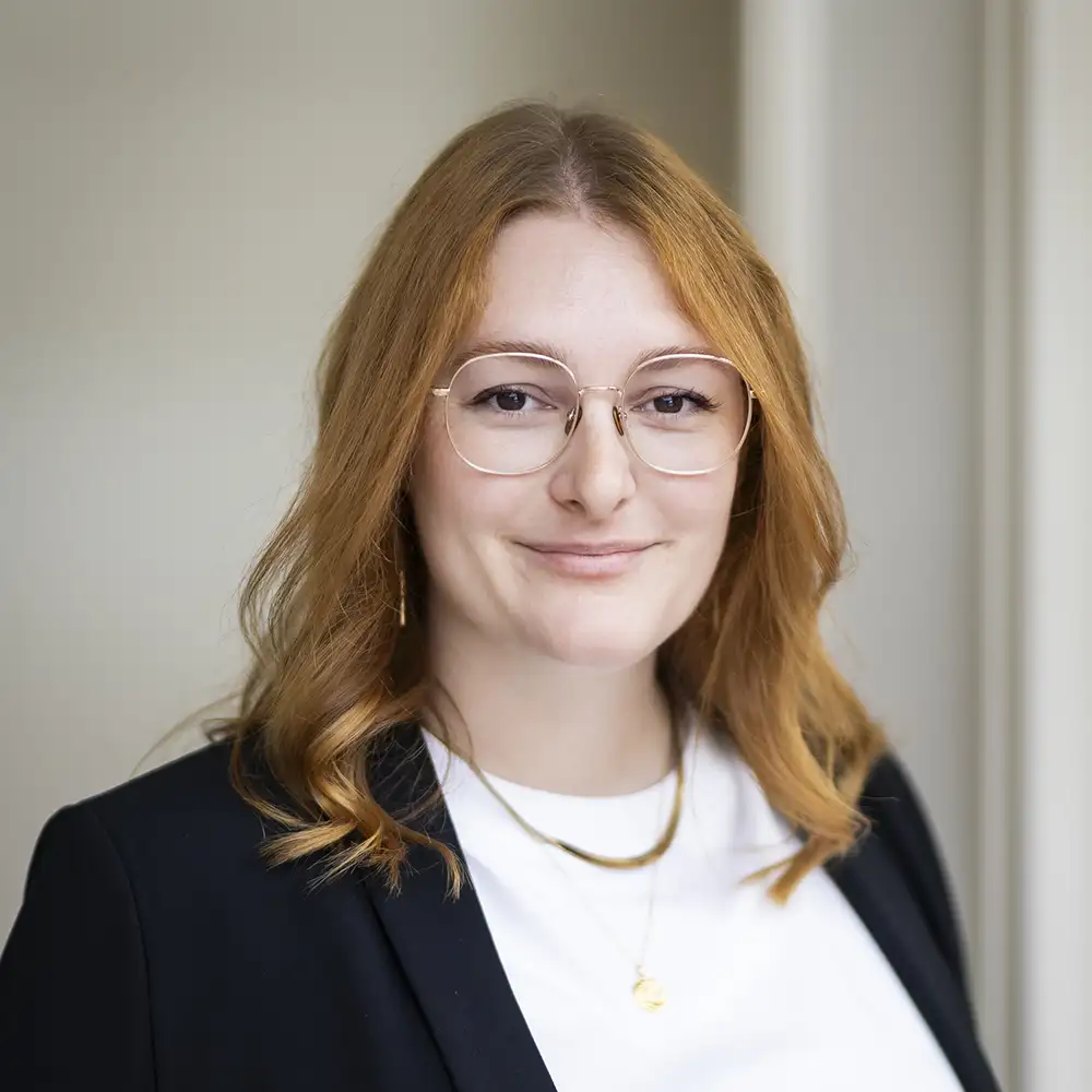 Une personne aux cheveux roux porte des lunettes et un blazer noir sur un fond neutre.