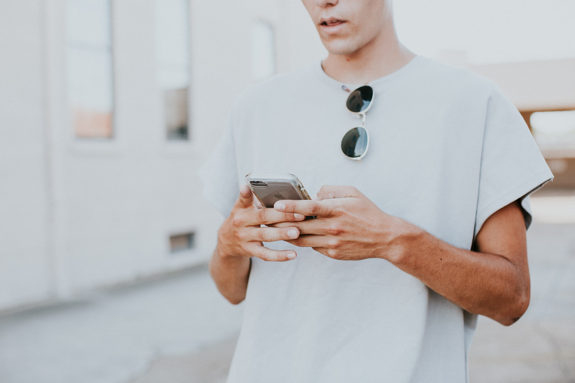 Une personne en t-shirt gris utilise un smartphone, avec des lunettes de soleil accrochées à son col, en extérieur.