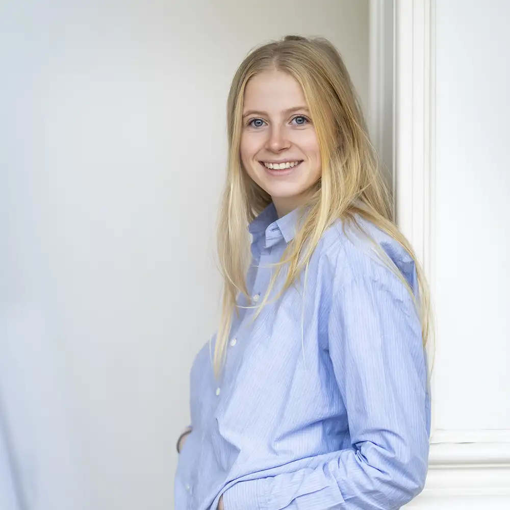Jeune femme aux cheveux blonds portant une chemise bleue, appuyée contre un mur blanc, souriant à la caméra.