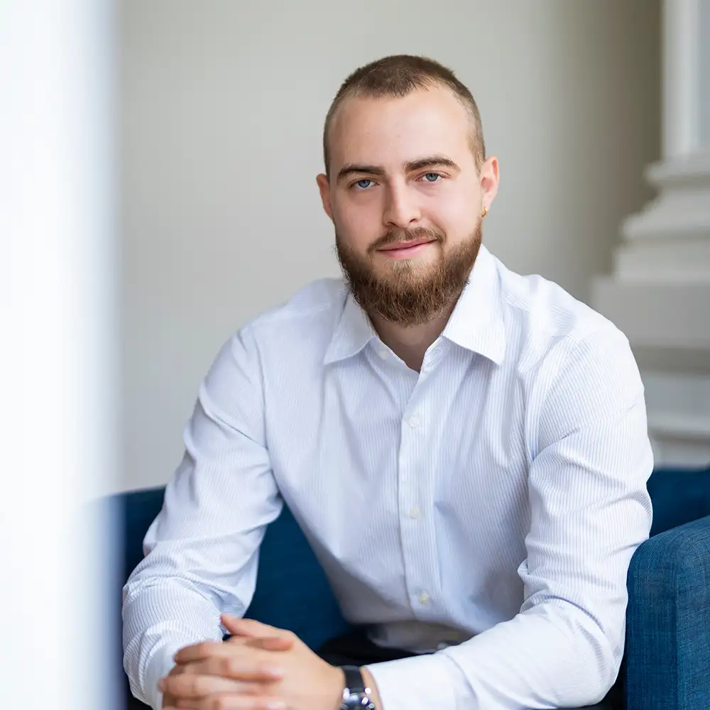 Un homme barbu en chemise blanche est assis sur un canapé bleu, mains jointes, fond blanc.