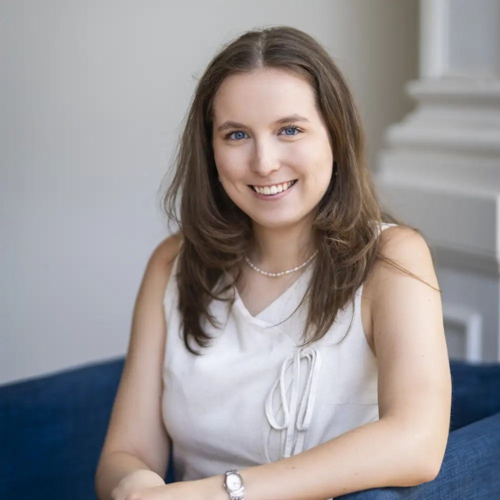 Une personne aux cheveux bruns porte un haut blanc et un collier, assise sur un canapé bleu devant un mur gris clair.