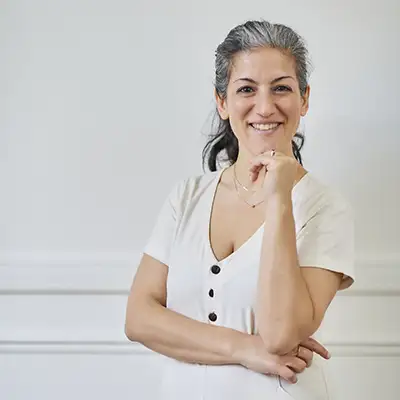 Une personne souriante aux cheveux gris et noirs, portant une robe blanche avec des boutons noirs, se tient devant un mur blanc.