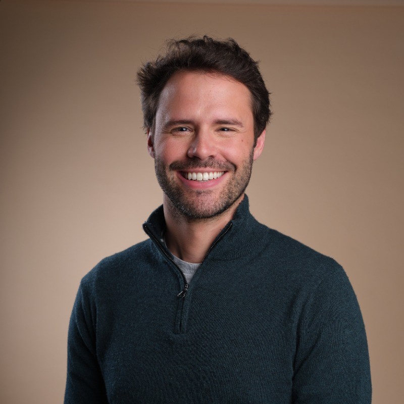 Un homme souriant porte un pull bleu foncé sur un fond uni beige.